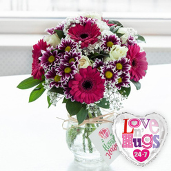 On a bright, uncluttered white surface, a vivid bouquet glows as if freshly arranged in a Clerkenwell studio, the kind you might find just off St John Street on your way toward Farringdon. The eye is first drawn to several generous gerbera daisies, their petals saturated in deep fuchsia and magenta, curling outward from dark, dramatic centres that add depth and contrast. Clustered among them are compact chrysanthemums, each little flower a study in detail, with rings of purple petals that gradually soften to pure white tips, circling a sunny yellow core. Nestled quietly within this lively mix, creamy white roses bring softness; their layered petals are still slightly cupped, hinting at the rose's familiar, gentle fragrance. Delicate branches of white baby's breath meander through the display like airy lace, lightening the arrangement and softening the transitions between the bold colours. The flowers and lush green foliage spill together into a clear glass vase, rounded and sturdy, its transparency revealing carefully trimmed stems standing in clear water. A length of rustic, natural twine wraps the neck of the vase, tied with an easy bow and carrying a small white tag, decorated with a simple pink rose motif and relaxed green writing, suggesting a hand-crafted, personal touch. Just to the right of the vase, a gleaming, heart-shaped foil balloon adds to the sense of celebration, edged in reflective silver and printed with a white centre where the words "Love n' Hugs 24-7" appear in playful patches of patterned pink, purple, and blue, sprinkled with little hearts. The soft, bright background hints at a window overlooking Clerkenwell's townhouses, giving the image a gentle, uplifting glow that suits anniversaries, Valentine's surprises, or an affectionate same-day delivery around EC1.