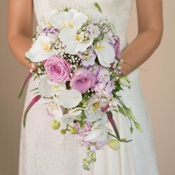 Bathed in gentle, natural light against a muted Clerkenwell backdrop, a bride is seen from the waist up, holding an exuberant cascading bridal bouquet that becomes an art piece in itself. The arrangement flows with drama and softness, featuring bold ivory phalaenopsis orchids that arch gracefully downward, their broad petals displaying a subtle shimmer and ochre centres. Full heads of blush pink and lavender roses create clouds of colour and fragrance; their velvety petals suggest the hands of a skilled local florist. Delicate white gypsophila tucks itself between these blooms, an airy finish reminiscent of Clerkenwell's hidden garden courtyards. Soft lilac and pale mauve blooms, likely sweet pea or stock, intermingle as textured accents. Below, vivid magenta strands of amaranthus sweep in rich, tactile ribbons, their texture lending modern drama to the bouquet's classic shape-an echo of Clerkenwell's blend of tradition and contemporary flair. Greenery and unopened orchid buds peek out, infusing the arrangement with freshness and movement. The trailing florals form an elegant waterfall effect, matching the gentle lacework of the bride's ivory gown glimpsed beneath her bouquet. The entire palette-muted whites, soft pinks, and tranquil lavenders-offers an air of romance and calm anticipation, ideal for a spring or summer ceremony. Crafted for local weddings by florists who know the neighbourhood's bespoke tastes, this bouquet stands as a stunning example of wedding artistry available in Clerkenwell.