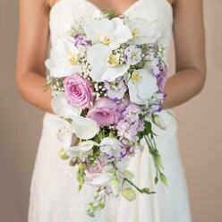 This evocative image captures a bride in Clerkenwell, mid-waist up, her posture poised yet gentle as she holds a lush, cascading bridal bouquet. The bouquet itself is a masterclass in textural and tonal harmony-a flowing teardrop design that bridges pure white, blush pink, and soft lavender hues, evoking the serenity of nearby Spa Fields on a tranquil morning. Striking phalaenopsis orchids reign at the heart, their deep yellow centers gleaming like sunrise over Charterhouse Square, while billowing pink garden roses provide warmth and classic depth. Fluttering between, clusters of airy lavender stock and white lisianthus-some edged with purple-create both visual movement and tactile softness, hallmarks of Clerkenwell Flowers craftsmanship. Accents of baby's breath and wisps of foliage weave in a natural, organic rhythm, giving the arrangement an ethereal, cloud-like presence. The strapless gown's intricate lace glimmers subtly at the bouquet's base, lending an elevated sense of bridal sophistication. There is a gentle fragrance imagined in the air-a mingling of orchard and wildflower-reflective of a wedding morning in London. Poised for a ceremony near St John's Gate or a modern loft in Clerkenwell, this bouquet radiates opulence and timeless romance, expertly constructed for couples seeking memorable wedding flowers intimately tied to the local spirit.