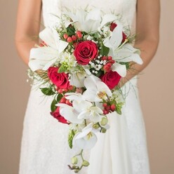 Captured in a luminous, painterly manner, this image features a bride dressed in a timeless white lace gown, gently holding an exquisite cascading bouquet designed with meticulous detail by a local Clerkenwell florist. The bouquet draws the eye with its prominent white oriental lilies, their silky petals and stamens opening amidst a cascade of gracefully trailing orchids. The deep red roses woven throughout exude classic romance, while vibrant hypericum berries punctuate the composition with festive splashes of colour. Interspersed sprigs of baby's breath and wispy green ferns create a sense of lightness, evoking memories of Clerkenwell's leafy squares and Victorian gardens. The sophisticated use of textures-from the velvet softness of rose petals to the crisp vibrancy of orchid blooms-feels both contemporary and steeped in tradition. A softly blurred, beige-toned backdrop ensures the vivid floral details and the bride's serene gesture remain central, making this bouquet an exceptional choice for a wedding in Clerkenwell, where historic charm meets modern artistry.