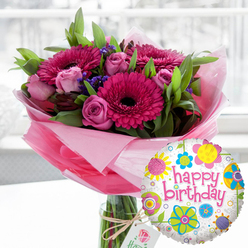 A joyful birthday bouquet and balloon pairing sits centre stage, captured in the kind of soft daylight you might find drifting through a Clerkenwell warehouse apartment window. In a tall, clear glass cylindrical vase, an abundant bouquet overflows with shades of pink and magenta. Large gerbera daisies in vivid fuchsia, each with a dramatic near-black centre, form bold focal points around which the rest of the flowers gather. Between them, blush pink roses and slightly deeper magenta rosebuds are scattered, some still tightly furled, others loosening their petals as if just beginning to perfume the room. Fine clusters of deep purple filler blooms, likely statice, are tucked throughout the arrangement, adding texture and a richer tonal layer, while lush, bright green foliage fans out to create a generous, rounded shape. The stems are embraced by soft, crinkled light pink cellophane or tissue that wraps the base of the bouquet inside the vase, adding a delicate, almost cloud-like effect. Around the lip of the vase, a length of rustic twine is tied into a casual bow, suspending a small, creamy gift tag adorned with a subtle rose emblem that hints at a luxury florist's branding. In the foreground to the right, a round foil "Happy Birthday" balloon leans playfully into the frame. Its white surface is sprinkled with tiny grey speckles and decorated with bright, whimsical floral and circular motifs in yellow, sky blue, lime green, pink, and purple, encircling the words "happy birthday" in bold pink script. The softly blurred interior background suggests a calm, modern London home, while the composition as a whole radiates warmth, celebration, and the simple pleasure of receiving fresh birthday flowers delivered locally around Clerkenwell.