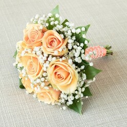 Cradled against the textured softness of a pale beige linen-like the kind often seen in Clerkenwell's artisan cafes-a bridal bouquet is artfully revealed in serene, even light. It centres around seven to nine plush, fully bloomed roses, their petals painted in a gradient from delicate apricot to sun-warmed peach, each spiral opening as if anticipating a gentle touch. Wisps of white baby's breath (Gypsophila) weave between the roses, their feathery clusters adding an almost cloudlike softness that heightens the bouquet's dreamlike charm. Peeking from beneath, rich green rose leaves provide a grounding flourish, their glossy hues evoking the leaf-dappled corners of Spa Fields. The arrangement, close and rounded, is bound into a classic spiral shape-its stem dressed elegantly with a peach-toned satin ribbon that glimmers with small white pearls, each one hand-positioned in a subtle spiral. The bouquet's base peeks out, freshly trimmed, suggesting it was arranged only moments before by a florist well-versed in Clerkenwell's vibrant wedding scene. The image is softly illuminated, capturing the subtle textures of petal, ribbon, and pearl alike, inviting admiration for every artful detail. Ready to grace a romantic ceremony from Farringdon to St. James's Walk, this design by Clerkenwell Flowers gently offers the promise of a love that blooms quietly yet endures, perfect for celebrations where elegance and heartfelt joy are prized.