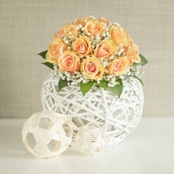 An elegant floral scene captures a rounded bouquet of soft peach and apricot roses, gathered so closely that they form a smooth, domed canopy of petals. Each rose reveals a gradient of colour, from a deeper, glowing orange core to paler, almost blush-toned outer petals, creating a warm, candlelit effect without any actual flame. Threaded delicately throughout the cluster, fine sprays of white baby's breath add a misty, cloud-like texture, their tiny blossoms softening the lines of the arrangement and enhancing its sense of lightness. Just below the main blooms, glimpses of deep green foliage frame the roses, giving the composition structure and the impression of freshly cut garden stems. The flowers sit in a striking white spherical vase, crafted from interlaced bands of material that form an open lattice reminiscent of woven rattan or sculpted string art. This airy structure allows light to pass through, creating intriguing shadows and a modern, sculptural silhouette you might expect to see in a design-focused home off Clerkenwell Road. Resting on the white tabletop in front of the vase are two decorative spheres in complementary pale tones, their woven, geometric patterns echoing the texture of the main container. One is slightly larger, one smaller, arranged casually yet thoughtfully, like styling details in a well-curated Clerkenwell loft. The background wall is a soft, neutral beige or greige with gentle vertical texture, keeping the focus on the flowers and their intricate vessel. The whole composition is bathed in soft, even light, evoking a feeling of calm celebration and understated luxury, ideal for a wedding centerpiece, a stylish dinner party, or refined gift delivery within the local neighbourhood.
