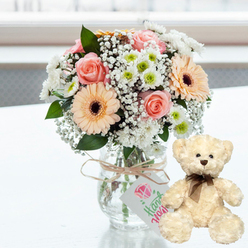 On a clean, white surface bathed in bright yet gentle natural light, a romantic anniversary tableau is carefully composed, reminiscent of a sunlit flat just off Clerkenwell Road. At the centre rests a clear glass jar-style vase, its transparency revealing a tangle of fresh green stems anchored in water. The vase cradles a lush, rounded bouquet of soft pink roses with velvety petals, pastel peach gerbera daisies with distinctive chocolate-brown centres, crisply petalled white chrysanthemums tinged with lime at their cores, and airy clusters of white baby's breath that soften the whole arrangement. Around the neck of the vase, a length of natural jute twine is tied in a casual bow, from which hangs a small white gift tag bearing a pink heart illustration and the word "Handmade" in elegant green script, adding a personal, crafted feel that suits the creative character of Clerkenwell's design studios. Just to the right of the flowers, a small cream-coloured teddy bear sits upright, its plush fur appearing irresistibly soft, with dark brown bead eyes, a matching button nose, and a slim brown satin ribbon neatly knotted at the throat. The bear faces slightly inwards, as if keeping the bouquet company, radiating comfort and gentle companionship. Behind them, the background fades into a bright blur, hinting at a window or light-filled space without drawing focus away from the gift set. This serene, tender composition captures the emotion of an anniversary surprise delivered across Clerkenwell, blending fragrant flowers and a cuddly keepsake into a single, heartfelt gesture.