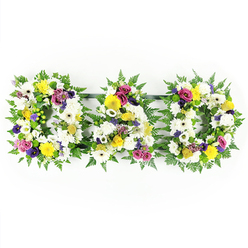 A striking "DAD" funeral tribute is carefully arranged in three separate block letters, photographed against a bright, uncluttered white background so every detail of the floristry stands out. Each letter is built on a sturdy frame and outlined in a generous collar of dark, glossy ferns and other lush green foliage, giving the design a soft, natural silhouette that feels at home in central London, much like the greenery tucked around Clerkenwell's historic streets and churchyards. Inside this leafy border, the florist has packed the flowers very closely together, creating a solid field of petals with no empty spaces. The dominant colour is white: large chrysanthemum heads and wide-petalled white gerbera daisies form a peaceful, traditional base, their layered textures suggesting comfort and serenity. Scattered among these are bright yellow gerberas, petite yellow spray roses, and small button chrysanthemums in warm golden tones, bringing gentle warmth and a sense of quiet optimism. Rich purple lisianthus blooms and clusters of magenta or deep pink spray roses add depth and emotional weight, while small tufts of lavender-coloured filler flowers and bright green button mums offer subtle contrast and freshness. All the flowers are turned to face front, ensuring the word "DAD" is bold and legible, whether resting by the coffin in a Clerkenwell funeral home or placed by the lectern for a memorial gathering nearby. This thoughtfully composed tribute captures both the dignity of loss and the tenderness of family memories, expressing love and remembrance in a clear, heartfelt way.