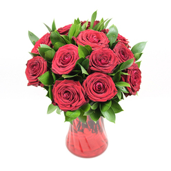 In this detailed studio image, a classic red rose bouquet sits proudly at the centre, framed only by a bright white background that lets every detail stand out in sharp relief. Roughly twelve to fifteen red roses form a rounded, compact cluster, their blooms at that perfect stage where the petals are fully unfurled yet still hold a sense of promise. Each flower reveals a tight spiral of velvety petals, creating a layered, almost sculptural surface that feels both soft and luxurious. The red is rich and deep, closer to wine or garnet than bright cherry, conveying warmth, devotion, and romance. Around and between the roses, a collar of fresh dark green foliage, likely ruscus, extends outward, adding volume and an organic, garden-like character. The leaves are smooth, slightly pointed, and glossy, catching the light and introducing cooler green tones that make the roses appear even more vivid. All of this is gathered into a translucent red glass vase with a gently curving body that narrows toward the base before opening out again at the rim. The glass has a faintly ribbed or undulating texture, so highlights dance along its surface, echoing the gentle twists of the rose petals above. The stems inside are hazy but visible, reinforcing the honest, natural feel. The bouquet radiates the kind of sincere affection you might send from an office off Clerkenwell Road to a loved one at home near St John's Square, a meaningful floral gift suitable for anniversaries, romantic milestones, or simply reminding someone how cherished they are.