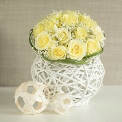 On a smooth white table, a luxurious dome of pale yellow roses glows softly, like early sunshine spilling over the terraces near Exmouth Market in Clerkenwell. Each rose is fully open, petals layered in generous swirls, their buttery tone made even gentler by the fine white mist of baby's breath threaded through the arrangement. These tiny gypsophila blooms sit like a cloud around the roses, lending them an airy, romantic halo that suits a wedding breakfast or elegant city reception. At the base, a narrow collar of glossy green foliage encircles the stems, defining the transition from lush flowers to the striking vessel beneath. The bouquet rises from a sculptural white woven vase, its openwork pattern of interlaced strands suggesting modern wicker, with gaps that play with light and shadow and give the impression the flowers are almost hovering above the table. To one side, two decorative spheres made from slender, light-toned strips of rattan or similar material echo the vase's texture, their geometric tangle of lines adding a gentle, contemporary twist. Behind, a softly blurred beige wall provides a calm, neutral backdrop, the kind you might find in a minimalist Clerkenwell studio, keeping all focus on the flowers and their craftsmanship. The overall mood is one of understated celebration and thoughtful design, ideal for a wedding table arrangement, an intimate anniversary dinner near St John's Square, or a refined centerpiece delivered by a local florist to bring soft colour and quiet happiness to a modern London home.