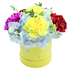 Seen from a slightly elevated angle on a neutral white field, this round floral gift box presents an expertly curated medley of colour and texture that reads like a small celebration. At the centre, two plump lemon-yellow carnations with ruffled edges anchor the design, their soft, almost satin-like petals layered to create a luminous core. Surrounding them are waves of soft periwinkle-blue hydrangea; the clusters of tiny florets form a cushiony base that both cools and amplifies the yellows' brightness. A vivid magenta carnation sits to the left, its rich tone introducing a lively contrast, while a velvety burgundy-red carnation on the right adds depth and a graceful counterpoint. Fresh green foliage threads through the bouquet, its fine stems and leaves giving the arrangement a natural, breathable silhouette. The box itself-pale lemon-yellow with scattered, irregular polka-dots in light blue, gentle pink and mint-feels whimsical; some spots carry minute white specks that catch the eye like confetti. As a Clerkenwell florist I note how the piece translates well for neighbourhood occasions: a birthday delivery to a Leather Lane studio, a cheery table accent for a Spa Fields gathering, or a thoughtful thank-you for a friend nearby. The flowers appear notably fresh, their textures ranging from the hydrangea's soft, papery florets to the carnations' layered frill, and the arrangement exudes a bright, uplifting energy. Clerkenwell Flowers prepares such boxed bouquets with attention to colour harmony and clean presentation, ideal for sending small moments of joy around Farringdon and the surrounding streets.