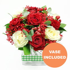 The image shows a richly detailed floral arrangement set against a clean white backdrop, the kind of bouquet you might see glowing in the window of a Clerkenwell side street florist on a crisp evening. At its centre, abundant deep red roses, fully open and velvety, are paired with softly glowing white roses, their petals elegantly curled and layered, like satin folded upon itself. This romantic red-and-cream palette is gently broken up by sprays of tiny red filler flowers, similar to coloured baby's breath, which form a misty halo between the larger blooms. Nestled among them are plump red hypericum berries, each one round and polished, catching the light and lending a festive, almost jewel-like quality to the piece. Fresh greenery, including eucalyptus leaves with their signature cool tone and rounded shape, threads through the bouquet, creating a sense of fullness and natural movement, as though the flowers have just been gathered from a market stall near Clerkenwell Green. The entire composition is arranged in a sleek, clear rectangular glass vase, the straight edges giving a contemporary balance to the soft curves of the petals. A green and white checkered gingham ribbon is wrapped neatly around the middle of the vase, evoking cosy kitchens, local cafés, and relaxed gatherings along St John Street. In the lower right corner of the photograph, a bold orange circle with white text clearly reads "VASE INCLUDED," highlighting that the design arrives as shown, ready to be placed on a dining table, office reception, or bedside, perfect for birthdays, anniversaries, or quiet at-home celebrations.
