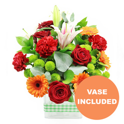 A lively mixed bouquet sits proudly against a pure white backdrop, presented in a modern, clear glass vase encircled at the base with a neat band of fresh light green and white gingham ribbon, reminiscent of picnic blankets in nearby Spa Fields. The flowers create a joyous, almost celebratory composition: velvety deep red roses and frilled red-orange carnations nestle side by side, their plush petals catching the light and giving the centre of the arrangement a warm, romantic glow. Bright orange gerbera daisies with dark, well-defined centres fan out around them, their flat, daisy-shaped faces adding a burst of optimism that feels perfectly in tune with Clerkenwell's creative energy. Scattered through the design are tight clusters of bright lime-green button chrysanthemums, their tiny, rounded heads adding a fresh, tactile contrast, like beads of colour. At the back and rising through the middle, elegant white Oriental lilies with soft pink accents lift the eye, some fully open with pollen-tipped stamens on display, others still wrapped in smooth buds, promising a slow, graceful unfurling over several days. The whole bouquet is framed with lush deep green foliage and long, slender stems of silvery eucalyptus that soften the outlines and add subtle movement, as though caught in a gentle breeze drifting up from Farringdon Road. In the lower right of the image, a round orange graphic partially overlaps the vase, clearly printed with the words VASE INCLUDED in bold white capitals, confirming that this contemporary glass container is part of the arrangement-ideal for quick, ready-to-display flower delivery to Clerkenwell apartments, studios, and offices.