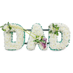 A solemn and lovingly detailed funeral floral tribute spells out "DAD" in three-dimensional letters, carefully arranged as though ready to be placed beside a coffin or memory table in a Clerkenwell chapel. Each letter is entirely covered with tightly clustered white chrysanthemums, or similar small white button blooms, forming a plush, snowy surface that looks soft to the touch and symbolises purity, respect, and a gentle farewell. Around the perimeter of each letter, a vivid emerald green satin ribbon is pleated into a ruffled border, neatly framing the snowy flowers and offering a fresh contrast reminiscent of the green spaces dotted around Clerkenwell, from Spa Fields to St James's Church garden. Behind the flowers, slim dark bars of the supporting frame peek through, revealing that the tribute is mounted to stand upright as one continuous display. The first "D" is embellished on its upper left curve with a small, considered cluster of white freesia blooms nestled into rich, glossy foliage, the stems arranged to look naturally grown rather than forced. Two slender, arching green stems extend from this area, sweeping diagonally across towards the central "A", where they open out into a striking yet understated spray. Here, two deep purple calla lilies, with their smooth, sculpted trumpets, rest among more white freesia flowers and vibrant green leaves, introducing a ribbon of colour that feels both regal and restrained. The final "D" carries a complementary spray of freesias and greenery at its upper right, slightly more compact but perfectly balanced. Set against a bright white background, this tribute conveys a moving sense of gratitude, loss, and enduring love for a father, ideal for memorial services and funeral ceremonies in Clerkenwell and the surrounding City streets.