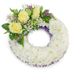 Presented on a stark white background, this circular sympathy wreath offers a composed, comforting presence formed from a plush mass of immaculate white chrysanthemums; their many-layered, ruffled petals create a tactile, cloud-like surface that reads as peaceful and consoling. A subtle touch of deep purple mesh or ribbon tucks through the inner and outer edges, visible in delicate glimpses, adding quiet depth to the primarily white surface. Positioned at the upper-left arc, a considered spray of accent flowers becomes the wreath's gentle focal point: three soft, creamy-yellow roses open in layered blooms, evoking warmth and enduring friendship, while fine clusters of white baby's breath lend an airy, breath-like softness. Interspersed among them are small lavender-purple statice or limonium flowers whose papery texture and muted hue bring a restrained pop of colour and visual rhythm. Lush green foliage - both broad, smooth leaves and slender fern-like fronds - frames the cluster and offers fresh contrast, their cool scent mingling with the faint rose perfume. The overall effect is one of refined sympathy and careful craftsmanship: even spacing, balanced weight, and seamless transitions from the dense chrysanthemum body to the accent cluster. As a local piece suitable for memorial services in Clerkenwell or delivery to nearby Farringdon or Leather Lane chapels, this wreath exemplifies thoughtful funeral floristry; Clerkenwell Flowers prepares such arrangements with attention to freshness and respectful presentation, fitting for a remembrance service or graveside tribute.