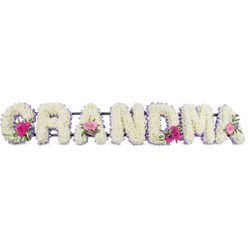 In this tender funeral flower tribute, the word "GRANDMA" is shaped from broad, plush letters, each one fully carpeted with compact white chrysanthemums that create a smooth, almost cloud-like surface. The blooms are tightly clustered, giving the impression of gentle strength and reliability, much like a grandmother's embrace remembered on quiet walks through Clerkenwell's side streets. A continuous lilac ribbon outlines every letter, its cool, satin sheen catching the light so that the edges seem to glow softly, adding refinement without overwhelming the calm white base. At three thoughtful points along the word, the white chrysanthemums part to reveal small, rounded sprays of mixed flowers. Pink gerberas with their open faces and tiny central discs sit alongside delicate pink roses, some in bud and some just unfurling, tucked amongst glossy green foliage that introduces movement and depth. These little islands of colour feel like treasured memories - bright moments within a life honoured with respect. The lighting is even and gentle, allowing each petal, leaf, and ribbon fold to be clearly defined, as if the tribute were resting in a peaceful Clerkenwell chapel or a quiet corner near Exmouth Market before a service. Though completely still, the design seems to breathe with emotion: a loving statement created with care by skilled hands, ideal for families in Clerkenwell wishing to say goodbye with grace. It is a floral arrangement that balances purity, softness, and personality in a single, meaningful word.