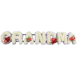 The image shows a beautifully composed funeral flower tribute spelling out GRANDMA in large, bold capital letters, laid out in a steady horizontal line against a plain, white studio-style background. Each letter is sculpted from tightly packed white cushion chrysanthemums or similar small-headed blooms, their rounded petals creating a soft, almost velvety texture across the surface. Tracing the edges of every letter is a subtle trim of pale purple ribbon or backing, giving a slight lavender haze that separates the crisp white flowers from the space around them and lends a sense of tranquillity. Along the tribute, vivid red accents are thoughtfully placed: the G holds a lush cluster of deep red roses and a distinctive red-and-white calla lily, nestled into one side like a cherished memory. The first A is gently marked with a single red anthurium, its glossy, heart-shaped spathe symbolising enduring love. At the centre, the M carries another grouping of red roses and a companion anthurium, rich in colour and emotion, while the last A echoes that sentiment with one final red anthurium at its base. The letters rest on a dark green metal or foam frame, mostly hidden, but strong enough to keep the tribute upright for display at a service. Crafted with the kind of detail you'd expect from an experienced Clerkenwell florist, perhaps preparing flowers for a farewell at nearby churches or crematoriums serving Farringdon and Bloomsbury, this personalised sympathy arrangement feels both respectful and deeply affectionate, offering a clear, loving farewell to a beloved grandmother.