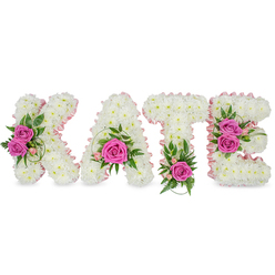 Handcrafted with careful attention to form and feeling, this three-dimensional floral name tribute spells out KATE in bold letters made entirely from compact, pristine white chrysanthemums. Each bloom is pressed closely to its neighbour to produce a seamless, cloud-like surface, the edges of the letters softly defined by a narrow pale pink satin ribbon that traces their interior curves. Bright hot-pink roses act as thoughtful focal points: the K bears two graceful roses on its left, the A cradles a single full rose at its lower centre, the T is punctuated by a solitary hot-pink bloom at its base, and the E carries a pair of roses along its right side. Complementary greenery - glossy ruscus, ferny fronds and a few variegated leaves - nestles around the roses, while tiny light-pink buds and delicate spray flowers add a whisper of softness. Subtle shadows beneath the letters indicate they are freestanding and substantial, ideal for laying beside a coffin or displaying on a memorial plinth. The overall impression is serene and affectionate: luminous whites counterpointed by compassionate pinks, suggesting the gentle fragrance of roses and the tactile richness of chrysanthemums. As a local florist in Clerkenwell, Clerkenwell Flowers often prepares arrangements like this for services at St John's Church, nearby Exmouth Market venues, and funerals in Farringdon; the piece conveys deep respect, personalisation, and a quiet celebration of a life.