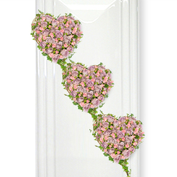 Three intertwined hearts made entirely of roses and ivy form a poetic funeral tribute, suspended visually against a spotless white background that lets every detail stand out. The hearts descend diagonally, from the upper left corner down towards the lower right, as if love itself were gently cascading down. Each heart is densely filled with miniature and spray roses, primarily in a soft, muted lavender that borders on dusty mauve, beautifully interwoven with clusters of pale blush pink blooms. The petals overlap to create a thick, cushion-like texture, almost cloud-soft to the eye, while a scattering of buds adds a sense of unfolding life and quiet hope. Fresh green ivy frames and connects the three hearts, its slender tendrils and polished leaves tracing each curve and then trailing elegantly onward, uniting the shapes into one flowing story. The foliage introduces a natural rhythm, balancing the romantic colours with a grounding, woodland freshness. To the left, faint vertical reflections suggest a modern, light-filled interior-perhaps reminiscent of a Clerkenwell gallery space or a serene chapel off Rosebery Avenue, where natural light pours in on days of remembrance. The overall arrangement feels calm, pure, and carefully considered, the work of an experienced local florist who understands how to express grief and gratitude through flowers. Ideal as a tribute at a funeral or memorial in and around Clerkenwell, this three-heart design speaks of enduring bonds, family ties, and the tenderness of a farewell offered with grace and love.