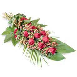 A long, flowing sympathy spray of red roses is displayed on a crisp white background, its diagonal placement drawing the eye from the neatly tied stems at the top left down to the graceful foliage that tapers at the lower right. Along the centre of the design, velvety red roses in different stages of opening are carefully placed so that buds, half-open blooms, and fuller flowers appear like a gentle wave of colour. Between them, clusters of small, coral-pink and pinkish-red Hypericum berries provide a soft contrast, their rounded forms and waxy surfaces echoing the curves of the rose petals while lightening the overall tone. The flowers sit on a rich bed of greenery: wide, dark green leaves with a glossy, almost lacquered finish create an elegant foundation, while finer fronds and leaf sprays, including bright, feathery palm leaves, fan outwards to create a sense of movement and length. The foliage feels both abundant and controlled, giving the tribute a sculpted, modern silhouette that would sit beautifully on a coffin or at the front of a memorial space. At the point where the stems gather, a smooth wrap of folded green leaf or ribbon holds everything in place, adding a sense of craftsmanship and care that reflects the work of an experienced local florist. This arrangement, which would be at home in a quiet Clerkenwell church or a City of London crematorium, visually conveys love, strength, and gratitude, making it a fitting final gesture for a farewell service or wake.