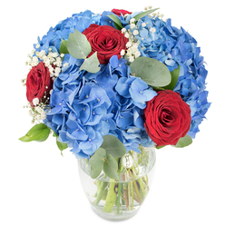 A carefully balanced floral composition in a clear glass vase, the arrangement reveals green stems immersed in water through its gently curved silhouette. Dominating the scene are voluminous clusters of azure-blue hydrangeas, their dense heads made of countless small petals that create a plush, cloud-like texture and a cool, saturated blue wash. Nestled among those blue masses are a few intensely red roses, their velvety petals tightly furled and offering a dramatic, romantic contrast that draws the eye. Scattered through the bouquet are soft white clouds of baby's breath, which lighten the composition and add a whispery, ethereal note. The inclusion of rounded eucalyptus leaves - their muted sage-green tones and smooth surfaces - introduces a subtle herbal aroma and a restful counterpoint to the saturated colours. The flowers are arranged with a confident, airy fullness that lets each element breathe while maintaining a cohesive, sculpted form. Bathed in bright, natural light, the colours read clean and saturated, making every petal and vein visible against the plain white backdrop. This is the sort of arrangement Clerkenwell Flowers might prepare for same-day pick-up or local delivery around Farringdon and Clerkenwell Green: striking enough for a milestone celebration, quietly romantic for an intimate dinner, or simply to bring a refined, patriotic colourway into a home on Charterhouse Street or nearby Leather Lane.