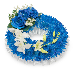 A thoughtfully constructed sympathy wreath photographed on a bright white background so every detail can be clearly perceived. The main ring is a lush, compact field of deep blue chrysanthemums whose countless tiny petals create a dense, tactile texture that reads like a soft carpet of colour. A neat band of pleated white satin lines the inner edge, its soft luster providing a delicate contrast and a refined outline to the vivid blue. Anchoring the design at the upper-left is a cluster of three fully opened blue roses, their petals rich and velvety in a striking azure tone; they are cradled by tiny white gypsophila stems and dark green foliage which add classic, comforting accents. Moving from the mid-left downward toward the lower right, a cascade of pure white orchids flows across the blue base, their broad, graceful petals lending an airy elegance and a sense of movement. Sprinkled along the orchid stem are fresh green buds in various stages of opening, quietly suggesting renewal and continuity. The mix of textures - the chrysanthemum's tight curls, the roses' velvet, the orchids' porcelain smoothness and the satin's crisp pleats - invites touch as well as sight. Bathed in soft natural-like light, the arrangement conveys solemn beauty and calm, appropriate for funerals or memorial services in Clerkenwell and nearby Farringdon. This is a tribute that reads as both dignified and tender, designed to express deep sympathy with understated refinement.