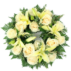 Presented from above on stark white, this funeral wreath is a study in gentle contrasts: a close-packed circle of creamy ivory roses, their layered petals curling inward like tiny, luminous shells, is threaded through with pale yellow trumpet lilies whose open blooms and sleeping buds create a rhythm of present bloom and quiet promise. The wreath sits on a rich base of dark green foliage - delicate fern fronds provide airiness at the edges while broader, rounded leaves akin to eucalyptus lend a softly matte, silvery cast that deepens the ivory and lemon tones. Approximately a dozen to twenty fully opened roses form the main visual field, their soft ivory catching light and softening the composition, while the lilies' elongated petals introduce vertical sweep and a restrained lemon warmth. The olfactory suggestion is subtle: powdery rose notes underscored by a green, almost citrus freshness from the lilies, a scent profile suited to memorial settings. The overall effect is carefully composed and comforting, the circular symmetry evoking eternity and unbroken remembrance. As a florist familiar with Clerkenwell and nearby Farringdon deliveries, Clerkenwell Flowers crafts this wreath with the measured restraint appropriate for a funeral or memorial, placing it gently at services around Exmouth Market, Leather Lane or St John's Square. It's designed to convey peaceful condolence, offering a serene visual tribute that reads as both dignified and tender.