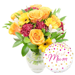 A bright, celebratory bouquet arranged in a simple clear glass vase sits against a pure white backdrop, a design that combines classic florist technique with an approachable warmth familiar to Clerkenwell clients. At the heart of the display are several large orange-leaning roses, their petals thick and softly spiralled, giving off a gentle, honeyed scent. Sunny yellow gerbera daisies peek between the roses, their broad faces and dark centres offering a sunshine-like punctuation that lifts the whole arrangement. Rich, deep maroon carnations add density and velvety shadow, their ruffled layers creating tactile depth beneath the lighter florals. Sprays of white alstroemeria, each petal dusted with faint speckles, introduce a delicate, airy note that catches the eye and softens transitions between colours. The greenery is abundant yet refined - glossy broad leaves and slim, graceful stems weave through the blooms and are visible through the vase, where the clean water and trimmed stems speak to the fresh-from-the-market care taken by the arranger. To the lower right the round white foil balloon reads "best Mum" in a flowing hot-pink script, its surface scattered with gold and bright pink polka dots and tiny hearts, lending a playful, affectionate accent that sits slightly in front of the vase. This composition reads as a confident gift choice for a beloved mother: warm, tactile and uplifting. As a local touch, Clerkenwell Flowers often pairs arrangements like this for deliveries around Leather Lane and Spa Fields, where the early light makes the orange and yellow tones sing.