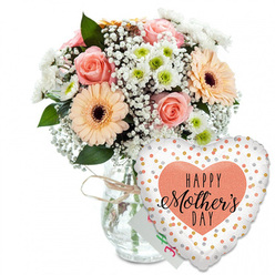 Set against a crisp white background, this Mother's Day scene captures the feeling of walking into a calm Clerkenwell studio filled with the scent of fresh blooms. At the heart of the image, a clear glass vase holds a generous, rounded bouquet, its transparency revealing clean stems neatly gathered by an experienced florist's hand. Soft peach-pink roses, fully open yet still tidy in their spiral, form the romantic core of the design. Beside them, bold peach gerbera daisies with deep chocolate-brown centres add a playful, almost sunny character, like a patch of light breaking through the clouds over nearby Exmouth Market. Between these focal blooms, clusters of delicate white gypsophila create a lace-like veil, while petite white chrysanthemums with light green centres add volume and a touch of botanical precision. The foliage, in various shades of fresh green, peeks strategically from behind petals and around the rim of the vase, giving the bouquet a natural, just-picked energy. Around the vase neck, a slim twist of rustic twine suggests a handcrafted, neighbourhood touch that feels typical of Clerkenwell's creative side streets. Tucked modestly behind the flowers is a small white card, its presence hinting at personal words of love rather than drawing attention away from the blossoms. In front and slightly to the right, a heart-shaped Mylar balloon rests against the arrangement, its white surface dotted with shimmering gold and silver circles. Centred on the balloon, a soft peach heart frames the message HAPPY Mother's DAY in bold black script, the sweeping letterforms mirroring the gentle curves of each petal. The entire composition evokes tender gratitude and quiet celebration, ideal for local Mother's Day deliveries around Clerkenwell and Farringdon.