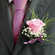 This image offers an intimate view of formalwear finished with a thoughtfully designed boutonniere, as though the wearer is about to step out onto one of Clerkenwell's cobbled side streets for a ceremony. The frame captures the torso from just below the shoulders down to mid-chest, showing a dark charcoal or nearly black suit jacket with a smooth, well-fitted lapel on the right side. A light-toned dress shirt, patterned with a fine, checked or gingham texture, peeks discreetly from beneath the jacket, giving visual interest without distraction. At the centre, a lustrous fuchsia-magenta tie, carefully knotted and pressed flat, draws the eye with its bold yet sophisticated sheen. On the left lapel rests a single pink rose boutonniere, clearly the focal point of the image. The rose is fully open but still fresh, its petals silky and layered, moving from a richer pink in the heart to a soft, blush-pink at the edges that almost glows against the dark cloth. Surrounding it, delicate sprays of white baby's breath cluster like airy mist, adding a gentle contrast and an almost lace-like texture. Fine, upright green foliage emerges from behind the blooms, echoing the look of slender grasses or herbs you might see in nearby Spa Fields on a bright day, bringing a natural, garden-picked feel. The stems are neatly bound with a deep green wrapping and finished with a slim, satin ribbon in mauve-lilac, tied into a neat little bow that sits just above the breast pocket. The ribbon's cool purple tone harmonises with the pink tie, creating a curated palette of charcoal, blush, white, and violet. The overall mood is tender and celebratory, ideal for weddings, civil ceremonies, or milestone events taking place in and around Clerkenwell, where every detail is chosen to mark a new beginning with quiet confidence.
