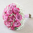A romantic rose bouquet rests at a graceful angle on a pale, textured cream backdrop, reminiscent of soft fabric draped in a Clerkenwell loft apartment. The arrangement is tightly composed, with about a dozen roses forming a rounded, hand-tied cluster. Each rose is a study in graduated colour: the inner petals glow with dusty mauve and lavender-pink tones, slowly deepening into vivid fuchsia and rich magenta at the fringes. The blooms are in various stages of openness, from firm buds to softly cupped flowers revealing intricate, spiralling hearts, giving the impression of quiet breathing and life. Enveloping the roses, a generous collar of white baby's breath creates a lacy, ethereal border, tiny blossoms hovering like a veil of mist. This gypsophila softens every edge, making the stronger pinks appear even richer while preserving an overall sense of lightness and air. Slender, deep green leaves hide shyly beneath the flowers, lending contrast without overwhelming the pastel palette. At the base, the stems are trimmed cleanly and bound with a sleek lilac satin ribbon, its sheen reflecting the soft daylight that might fall through Clerkenwell's large warehouse windows. A delicate strand of white pearls twines around the ribbon, catching the light and evoking bridal accessories and heirloom details. The image feels still and contemplative, as if the bouquet is pausing between being finished by an expert florist and being carried to a wedding near St John's Square. It suggests tender celebrations, civil ceremonies, engagement parties, and cherished anniversaries framed by simple, thoughtful design.