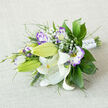 Set against a pale, woven fabric that recalls the soft neutrals of Clerkenwell loft apartments, this horizontal bouquet showcases the calm elegance of classic wedding florals. The eye is drawn first to the white Oriental lilies, some fully open with wide, curving petals that catch the light like satin, others still held in plump, green buds. Their prominent yellow-orange stamens are lightly dusted with deep brown pollen, and faint speckles trace the inner petals, adding refined detail. Nestled among the lilies, white lisianthus blossoms edged in rich purple create a beautiful contrast, their ruffled petals curling like brushstrokes of paint along the margins. Part-open lisianthus buds, tinged with violet at the tips, give the sense that the bouquet was freshly arranged that morning, perhaps just a short walk from St John Street or Farringdon. Misty clusters of baby's breath weave through the blooms, scattering delicate white stars that soften the composition and lend a cloud-like lightness. Around the edges, a generous selection of greenery provides structure and fullness: broad, glossy leaves, feathery fern-like sprigs, and slender grassy spikes all overlap to create tiers of textured green. The stems are neatly bound with a white satin ribbon, its surface dotted with tiny, pearl-style accents that feel quietly luxurious and bridal. Crafted with care in the style of an intimate wedding bouquet, this arrangement suggests purity, devotion, and a serene joy-perfect for ceremonies in Clerkenwell churches, registry offices, or chic warehouse venues, and ideal for same-day delivery when only something graceful and timeless will do.