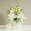 In this thoughtfully composed wedding arrangement, the gentle elegance of white and lilac blooms is showcased with the quiet confidence of an experienced Clerkenwell florist. Three or more open white lilies form the visual anchor, their broad, silky petals curling back to display softly speckled centers and prominent yellow-orange stamens that feel almost sunlit. Interlaced around them are lisianthus flowers in creamy white, some with petals kissed by a fine lavender edge, giving the impression of watercolour brushstrokes. Several pale green buds, both lily and lisianthus, rise from within the bouquet, like new stories beginning along the cobbled side streets off St John Street. Fine sprays of dark green foliage weave in and out, joined by lacy clusters of white filler flowers reminiscent of gypsophila and tiny deep blue or purple blossoms that punctuate the design with jewel-toned accents. All of this is cradled in a bulbous white vase crafted from tightly woven strands, its sculptural, basket-like texture catching the light and adding a modern, artisanal feel that would suit a loft venue near Farringdon. Resting on a flawless white surface, the vase is accompanied by two decorative spheres of pale rattan or bamboo, their natural weave echoing the vase and softening the overall look. The background is a muted beige with a delicate vertical woven pattern, suggesting a fabric panel or textured wall. Bathed in bright yet gentle studio lighting, the bouquet radiates calm celebration, making it perfect for wedding collections, registry office ceremonies, or intimate receptions across Clerkenwell, where understated beauty and thoughtful detail are treasured.