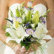 A refined bridal bouquet that balances classic formality with whisper-soft colour: large white lilies take centre stage, their broad, star-shaped petals unfolding with a creamy sheen that reflects gentle sunlight. Scattered between them are lisianthus blooms edged in a subtle violet, their ruffled petals offering a cool contrast and painterly texture. Along the stems, clouds of tiny baby's breath introduce a delicate, lace-like softness while slender blades of bright green foliage and delicate grass-like tendrils provide movement and an open, breathable frame. Small veronica spikes and a few tight buds add sculptural points and suggest promise, the way a gown might have hidden details revealed as you turn. The fragrance is floral and uplifting - the unmistakable sweetness of lilies tempered by fresh, green notes.

Crafted with the steady hands of a local florist, this bouquet carries the kind of quiet expertise you'd expect from a shop serving Clerkenwell and nearby Farringdon; at Clerkenwell Flowers we think of it alongside early-morning photographs on Exmouth Market or a reception near Smithfield's old arches. The arrangement is tied so each stem contributes to a unified, elegant silhouette, suitable for intimate city weddings and airy church ceremonies alike. It translates beautifully into pictures and memory: soft whites become luminous in daylight, violets read as gentle accents, and the overall composition feels both ceremonial and warmly personal - a carefully considered heirloom of the wedding day.
