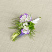 There is a tender language in this boutonniere that speaks of spring mornings and intimate vows: two lisianthus heads, white with a whisper of violet along their ruffled edges, form the focal point, their velvety petals folding inward like a small, sheltered world. Specks of baby's breath hover between stems like soft white constellations, giving the piece an airy, breathy cadence, while narrow clusters of grape hyacinth punctuate the lower edge with cool blue-violet spikes that add vivid contrast. Wisps of fern thread through the grouping, their feathered fronds suggesting movement and a woodland freshness that tempers the formal shape. The stems are gathered and tied with a crisp white satin ribbon, its smooth texture complementing the linen surface beneath, and the arrangement is lit so that the colours read cleanly against the neutral backdrop. Smell is subtle-a faint green, slightly sweet fragrance-while touch reveals silk-soft petals and the coaxing sturdiness of the stems. Picture this boutonniere pinned for a small Clerkenwell wedding, perhaps after a ceremony near Farringdon Station or a celebratory portrait on Cowcross Street; it fits both modern tailoring and classic suits. As a florist who works locally, I craft these with attention to how they sit on fabric, how they hold through a long afternoon, and how they quietly complete a look without shouting. It's a detail that leaves an impression precisely because it is composed with restraint and sincere care.