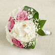Laid gently on a pale, textured fabric that looks like natural linen in warm beige and ivory tones, this bouquet has the unhurried elegance of a Clerkenwell morning before the streets grow busy. At its centre, a generous creamy white peony blooms in layered, feathery petals, each fold catching the soft, diffused light like brushed satin. Around it, vibrant fuchsia-pink roses cluster in various stages of opening, their cupped, spiralling forms adding structure and a pulse of romantic colour, as if ready to be carried from a studio near St John Street to a wedding at a hidden courtyard venue. Wisps of white gypsophila thread between the larger flowers, hundreds of tiny blossoms forming a cloud-like halo that lightens the composition and adds delicate texture. The greenery is lush and assured: broad, deep green leaves with a natural gloss, some arching protectively around the blooms, others peeking out from beneath, echoing the leafy calm of nearby Spa Fields. Each stem is carefully gathered and bound with a pristine white satin ribbon, the wrapping smooth, secure, and subtly reflective, creating a tidy, comfortable grip that reveals the hand of an experienced florist. Shadows are soft and understated, helping each blossom stand out without drama, and the overall impression is one of thoughtful craftsmanship and quiet joy. This wedding bouquet combines creamy whites, vivid pinks, and fresh greens into a design that feels both timeless and contemporary, ideal for ceremonies and celebrations throughout Clerkenwell and the surrounding central London streets.