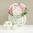 A lush, dome-shaped bouquet of white peonies and vivid fuchsia-pink roses is showcased as though styled for a chic Clerkenwell wedding, its romance enhanced by soft, even lighting. The peonies sit at the heart of the arrangement, their ivory petals layered and slightly ruffled, like delicate silk folded again and again, suggesting a gentle, powdery scent. Around them, pink roses in full bloom offer velvety spirals of colour, glowing from blush to rich rose, creating a sense of warmth and joyful celebration. Wisps of baby's breath are threaded throughout, their fine, branching stems tipped with tiny, cloud-like flowers that lend an airy, floating quality to the design. Framing the blooms, strong, smooth green leaves arc outward, their rich colour and polished surface giving the bouquet a fresh-from-the-garden energy that feels right at home near Clerkenwell Green or the studios off St John Street. The flowers rest in a distinctive white, openwork vase, its woven, branch-like structure forming an airy cage that looks both handcrafted and contemporary, allowing hints of stems and shadow to peek through. Two matching woven spheres, light in colour and texture, sit nearby on a clean, white surface, echoing the vase's rhythmic pattern and adding a minimal, sculptural detail. A soft beige background, reminiscent of textured fabric or plaster, keeps the focus squarely on the interplay of petals, leaves, and weaving. The overall mood is serene, refined, and welcoming-perfect for a registry office bouquet, an elegant table piece for a Clerkenwell restaurant reception, or a thoughtful gift delivered locally to mark a milestone moment.