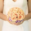 A close-up photograph captures a bride from the chest down, standing in soft, natural light that could easily be streaming through a Clerkenwell townhouse window before the ceremony. She wears a classic white wedding gown with an ivory undertone, the bodice gently shaped into a modest V-neckline and detailed with fine lace that traces delicate floral patterns across the fabric. The material around her waist falls in subtle folds, giving the dress a timeless, romantic structure. Cradled in both hands is a carefully crafted, round bridal bouquet, held at mid-torso so it becomes the quiet centre of the image. The bouquet is composed mainly of tightly clustered roses in warm, tender shades of peach, blush pink, and light apricot, their petals curling inward in soft spirals like shells. These roses are arranged in a domed shape, each bloom nestled closely to the next to create a sense of fullness and abundance without feeling heavy. Beneath this cloud of roses, a border of ruffled flowers in deep purple with white, almost feathered edges-likely lisianthus-forms a vivid ring that frames the arrangement and adds depth. Tiny white baby's breath is tucked here and there around the base, breaking up the colour and adding a misty, lacy texture. The bride's fingernails are neatly manicured in a pale, neutral polish, with one thumb showing a delicate hint of shimmer that catches the light. The overall mood is calm, poised romance, like a quiet moment on a side street near Exmouth Market, just before the celebrations begin-a bouquet that reflects the soft elegance many couples in Clerkenwell seek for their city weddings.