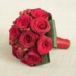 This exquisite Clerkenwell bridal bouquet showcases around ten opulent deep red roses, each one blooming in satiny swirls of rich colour, exuding both warmth and sophistication. The roses are gathered in a neat, rounded shape, their intense scarlet petals drawing admiring eyes and conveying a sense of passion. Interwoven throughout are glossy, bright red hypericum berries, their clustered forms and glistening surfaces standing out against the velvet smoothness of the flower heads. Framing the bouquet with care, broad, lush green leaves have been wrapped at the base for visual structure and contrast-an artistic nod to Clerkenwell's celebrated design sensibilities. The stems are tightly bound and finished with an elegant length of deep red satin ribbon, its sheen catching the soft, even light that fills the scene. The arrangement is set upon a subtly textured backdrop-perhaps reminiscent of fine linens found in local cafés near Leather Lane-allowing the vibrant reds and fresh greens to shine. The bouquet's composition speaks of a skilled florist's hand, its mood joyful and tender, ideal for wedding celebrations or anniversaries in the heart of Clerkenwell, where floral artistry and love go hand in hand.