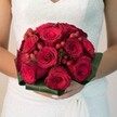 Nestled within the urban vibrance of Clerkenwell, this evocative bridal bouquet exudes romance and sophistication. The image, rendered in softly diffused daylight, centers on a stunning arrangement-a lush, rounded clutch of deep red roses whose petals swirl in layered spirals, alternating between youthful buds and sensuous, open blooms. Sparkling among the rich reds are petite crimson hypericum berries, their glossy finish giving the bouquet a modern, almost jewel-like flourish, reminiscent of a hidden garden courtyard near Leather Lane. Encircling the composition, broad, verdant leaves are artfully arranged in a neat collar, the subtle sheen of their waxy surfaces echoing the vitality of nearby Clerkenwell Green. The bride's gentle hands, manicured and poised with care, cradle the blooms lovingly above her vintage-inspired lace wedding gown, whose delicate floral motifs and flowing fabric are softly visible at the image's edge. The bouquet's vibrant palette radiates warmth and high emotion against the understated elegance of her dress, evocative of a day filled with whispered vows and joyful celebrations in local historic venues. Every detail-from the textural interplay of supple petals to the striking crimson berries-offers a tactile feast for the imagination, inviting thoughts of fleeting scents and the quiet thrill of a wedding ceremony close to home. This timeless arrangement, expertly crafted for weddings in Clerkenwell, speaks of promise, unity, and vibrant beginnings.