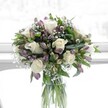 A softly lit floral arrangement stands in a clear, cylindrical glass vase, placed against a bright, gently diffused background that suggests a sunlit Clerkenwell window or pale studio wall. At the heart of the bouquet, several creamy white roses are fully open, their velvety petals curling outward in perfect spirals that convey quiet romance and refinement. Around them, clusters of lavender and muted purple alstroemeria, some still in tight bud and others just beginning to unfurl, add layers of delicate colour, their petals veined with deeper purple tones that reward a closer look. Airy white sprigs of baby's breath weave between the larger blooms like tiny clouds, softening the edges and lending an almost mist-like lightness to the overall composition. Lush greenery in varied shapes and textures-broad, dark leaves, slimmer lance-like foliage, and neat green stems-frames the flowers and provides structure, so the bouquet appears full and rounded without feeling heavy. Through the transparent glass vase, you can see the stems cleanly cut and arranged in fresh water, evidence of careful, professional conditioning. The palette of ivory, lilac, and cool green feels calm and restorative, reminiscent of an early morning walk through Spa Fields or a quiet corner near St John Street. The light appears natural, perhaps from a Clerkenwell studio window, giving the flowers a soft glow that enhances their natural beauty. This image evokes tender celebration and thoughtful care-a bouquet that might be delivered locally for a gentle get-well wish, an understated birthday, or a message of comfort and hope in the heart of EC1.