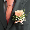 This boutonniere is composed with an eye for balance and intimate detail: a single peach rose sits serenely on the lapel, its velvety petals glowing with soft coral-pink warmth and edged in a creamy blush that catches the light. Around it, tiny clusters of white gypsophila create a cloud-like frame, airy and ethereal, while slender, fresh green foliage and a lightly curled supporting leaf add structural rhythm and a lively, garden-fresh note. The rose's subtle fragrance is suggested rather than proclaimed, a sweet, uplifting scent that mingles with the quiet fabric of the suit. A satin ribbon, matching the tie's coral hue, is tied cleanly at the base, echoing the careful handiwork of a florist and the personal touch of someone fastening the bloom for a beloved. Set against a charcoal jacket, every colour reads truer and more luminous; the deep grey acts as a calm canvas that heightens the boutonniere's gentle cheer. Visually, the composition balances tenderness with restraint, making it well-suited for ceremonies or milestone celebrations - the kind of small, meaningful piece you might see at a wedding near Exmouth Market or arriving with a thoughtful order across Clerkenwell and Farringdon. The overall feeling is quiet celebration: modest in size but rich in texture and sentiment, crafted to accompany vows, toasts, or an elegant evening out.