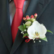 Pinned to the left side of a charcoal suit jacket, this boutonnière reads like a small, cultivated moment of celebration-one we often prepare at Clerkenwell Flowers for weddings and formal evenings around Exmouth Market and Clerkenwell Green. The outfit beneath the lapel reveals a soft, light-coloured dress shirt with a faint grid pattern and a bold, satin red tie whose chromatic intensity provides the visual anchor. Center stage is a single white Phalaenopsis orchid, its smooth, almost luminous petals unfurling in a calm, sculptural way; the orchid's labellum shows the signature yellow-gold markings that give the bloom a jewel-like heart. Surrounding the orchid, deep green glossy foliage shapes a steady backdrop, while wisps of gypsophila create a delicate halo of tiny white dots that soften the silhouette. A small cluster of red hypericum berries adds a cheerful, rounded punctuation that mirrors the tie's colour and introduces a subtle tactile contrast against the orchid's velvety petals. A thin red satin ribbon is tied at the stems, a neat finishing gesture that repeats the satin sheen and secures the stems to the lapel. The light in the photograph is gentle-like early evening by St. John's Square-rendering textures with quiet clarity: the cool silk of the tie, the waxy smoothness of the orchid, the fragile powder of baby's-breath blossoms. It feels bespoke and poised, ideal for marking vows, anniversaries, or a black-tie event in and around our neighbourhood.