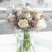 A tranquil bouquet of white roses and purple filler flowers rests in a clear glass vase, captured in soft, natural light that feels very much like a late morning in a Clerkenwell apartment overlooking Exmouth Market. The roses are fully opened, each bloom a careful spiral of creamy-white petals with the faintest suggestion of ivory at the core. They are not crowded, but comfortably nestled together, their rounded forms creating a gentle rhythm across the top of the arrangement. Threaded between them are feathery sprays of tiny lavender and pale pink blossoms, likely statice, forming a delicate haze around the more substantial roses. These miniature flowers add a veil of colour and light texture, softening the bouquet's profile and lending a quiet, dreamlike quality. The tall, cylindrical vase is perfectly clear, revealing straight, cut green stems descending into fresh, unclouded water; the stems cross in subtle, natural lines, giving the design an honest, uncontrived beauty. The vase stands on a pristine white surface that amplifies the brightness of the scene, while the background melts away into a softly blurred wash of pale tones. There are no distractions-only the flowers, the light, and the sense of stillness they create. The overall feeling is one of gentle condolence and tender remembrance, as if this bouquet has been chosen to sit on a side table in a quiet Clerkenwell home after a funeral service, offering comfort through its simplicity and calm. It's an image that speaks of respect, kindness, and the enduring solace of fresh flowers.