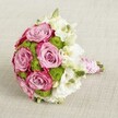 A luxurious bridal-style bouquet lies against a softly textured light beige fabric, photographed from a slightly elevated angle so the rounded shape appears full and perfectly composed. At the centre, several large, fully opened roses in soft lavender-pink draw the eye, each petal curling outward in velvety layers that deepen to a richer, almost fuchsia edge, creating a romantic two-tone effect. Nestled between these statement roses are neat clusters of bright chartreuse green button chrysanthemums, their tiny, domed heads adding a lively, modern contrast in both colour and texture. Around and between the main blooms, delicate white flowers, likely lisianthus, unfurl in ruffled, cloud-like layers with greenish-yellow centres that hint at gentle fragrance and absolute freshness. A few tight green buds peep through, suggesting the bouquet has been freshly arranged and will continue to open throughout the day. The stems are cleanly trimmed and tightly bound to form a smooth, hand-tied handle wrapped in pale pink satin ribbon that catches the light with a soft sheen. Over this ribbon, a single strand of small white pearls is wound in a graceful spiral, giving the handle the feel of heirloom jewellery and making the bouquet ideal for a stylish Clerkenwell wedding or civil ceremony near Exmouth Market or Spa Fields. The overall mood is one of refined celebration and tender emotion, perfect for a bride, a special anniversary, or a luxurious gift that mirrors the understated elegance often seen in central London gatherings.