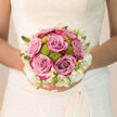 Held close to the bride's waist, this elegant wedding bouquet feels like a calm pause in the excitement of the day, its colours soft and reassuring. In the centre, a cluster of six roses in a gentle lilac-dusty pink tone open in perfect spirals, their petals layered like fine silk, recalling the refined textures found in Clerkenwell's design studios and historic townhouses. Nestled between them, bright lime-green button chrysanthemums create playful, rounded bursts of colour, almost like tiny lanterns of fresh spring energy glowing among the pastel blooms. Along the outer edge, small ivory and white blossoms, some just beginning to open from pointed buds, trace a delicate border that brings to mind intricate lace panels you might glimpse in a bridal window on nearby Farringdon Road. The bouquet rests against a white wedding dress with subtle texture, and the overall composition is gently rounded, balanced so that no single flower dominates, yet the roses clearly hold the emotional focus. Soft, natural light - the kind you might find streaming through a Clerkenwell loft window - slips across the petals, highlighting their gentle curves and casting barely-there shadows within the bouquet's depths. Imagining its scent, you might notice a blend of classic rose perfume with clean, green tones and a whisper of earthiness from the stems freshly trimmed that morning. The way the flowers sit so comfortably in the bride's hands suggests both structure and ease, a design created by an experienced local florist who understands how to craft a bouquet that feels intimate, modern, and ready to stay beautiful from ceremony to reception in Clerkenwell.