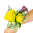 A compact, wearable work of floral jewellery rests diagonally across a light-to-medium forearm, photographed against a crisp white background so every detail reads clearly. Two buttery yellow roses form the joyful centre: one noticeably more open and ruffled on the left, the other a slightly cupped bud to its right, both with soft, fragrant petals that suggest the gentle scent of fresh roses. Each rose is given a small clear rhinestone pressed near its heart, the tiny crystals catching the studio light and lending a discreet sparkle when the wrist moves. Behind and above the roses a single, trumpet-shaped calla lily in an almost-black cherry plum unfurls with satin-smooth texture, its deep colour providing a sophisticated counterpoint to the sunny yellows. The composition is built on layers of greenery - fine, feathery asparagus fern that breathes airiness into the piece and broader variegated leaves with pale, buttered edges that feel cool and waxy to the touch. Little clusters of white button-like filler flowers nestle among the foliage, giving the corsage a sense of fullness and delicate detail. The band securing the arrangement is slim and mostly hidden beneath the flowers, so the design reads as an organic extension of the wrist, angled from the bottom-left of the frame toward the middle-right. Soft, even lighting highlights texture - the velvety rose petals, the calla's polished sheen, the fern's filigree - and the overall mood is celebratory yet refined, suitable for prom, a wedding party, or any formal moment when a wearer wants a polished flash of sunshine and evening plum at their sleeve.