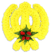 A substantial, sculptural tribute formed almost entirely from countless small, bright yellow chrysanthemum blooms creates a friendly, stylised silhouette against a clear white backdrop. The chrysanthemums are set tightly together so their rounded heads read as a single, plush surface; under soft studio light their petals pick up a warm golden sheen and the dense texture suggests a velvety cushion of colour. Rising on either side are two curved lobes that give the overall contour a playful, almost mascot-like quality, yet the craftsmanship remains precise and reverent. Anchoring the design at the bottom centre is a compact cluster of five rich, deep red roses, their velvety petals arranged in tight buds and hugged by deep green fern fronds that fan out to frame the reds. The contrast between the sunny yellow field and the concentrated crimson heart is immediate and deliberate-cheerful yet solemn, celebratory yet respectful. A faint grey shadow beneath the piece confirms it sits as a physical tribute on a tabletop or easel, intended for display at a memorial, themed celebration, or a personalised funeral arrangement. The scent is a soft crossover of green, slightly sweet chrysanthemum and the classic perfume of red roses, a quiet reminder of care in selecting each stem. Locally, this kind of bespoke display is one we would sketch for commissions around Exmouth Market or for a thoughtful offering at Clerkenwell Green, where neighbours often request symbolic, characterful tributes for community events or private commemorations.