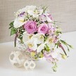 A sophisticated floral arrangement is presented in a close-up view, as if positioned along a wedding banquet table in Clerkenwell, its colours and textures carefully balanced to create a sense of refined celebration. The flowers are gathered in a round white wicker basket with a snug, handwoven pattern, the painted strands catching light and shadow to form a subtle, tactile backdrop for the blooms. Dominating the arrangement are several fully-opened pink roses, their petals forming intricate spirals that radiate outward from tight centres, suggesting romance, grace, and affection. Weaving gently among these roses are white Phalaenopsis orchids, their smooth, broad petals fanning out like wings around golden-yellow throats, bringing a touch of contemporary elegance that contrasts with the classic roses. Around them, soft white Lisianthus blossoms, some with delicate purple-tinted edges, introduce an extra layer of texture and gentle colour variation. Airy tufts of white baby's breath are interspersed between the larger flowers, creating a cloud-like effect that lightens the arrangement and draws the eye inward. Pale pink stock stems extend slightly beyond the main mass of blooms, their clustered florets cascading with a natural, almost garden-like movement. Glimpses of glossy green foliage and a few slim, trailing leaves add depth and freshness, hinting at the bouquet's just-cut quality from a local studio near Clerkenwell Green. The basket rests on a smooth white surface, beside two decorative spheres woven from pale natural fibres, their layered, looping strands echoing the basket's rustic charm and adding sculptural interest. Behind it all, a soft beige or taupe fabric background with fine, linen-like texture creates a warm, neutral stage that allows the pinks, whites, and greens to glow. The overall impression is one of quiet luxury and joyful romance, perfectly suited to wedding receptions, engagement dinners, or intimate celebrations across Clerkenwell.