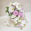 An elegant, cascading floral design is displayed on a smooth white tabletop, conveying the feeling of a wedding breakfast set in a stylish Clerkenwell venue off Old Street. The arrangement is gathered into a distinctive white spherical vase with an open woven lattice, like a decorative rattan ball transformed into a modern centerpiece. From this airy container, tall stems of white Phalaenopsis orchids rise and tumble, their velvety petals broad and pristine, with soft yellow and golden throats adding a hint of warmth and depth. Around the orchids, lush roses in shades of pastel pink and gentle lavender are fully opened, their many layers of petals curling outwards in soft spirals that suggest romance and tenderness. Smaller, frilled flowers, probably Lisianthus, contribute additional texture; their white petals brushed with lilac or purple on the edges create delicate strokes of colour among the paler blooms. Wisps of white baby's breath thread throughout like a fine, lacy mist, giving the composition a light, airy quality and helping each flower blend harmoniously with the next. From the lower edge of the arrangement, slender stems of light purple stock or similar trailing flowers flow downward in a soft cascade, hinting at the movement of a bridal bouquet draped over a table. Glossy green leaves and subtle foliage are tucked between blooms, adding contrast and a fresh, garden-like feel. On the white surface nearby, two pale woven spheres-one slightly larger, one smaller-mirror the texture of the vase and complete the tranquil vignette. Behind it all, a softly textured beige wall recedes gently out of focus, letting the soft colours, craftsmanship, and sense of celebration remain firmly in the spotlight.