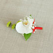 On a pale, linen-like background that recalls the softly worn textiles of a Clerkenwell loft, a sophisticated boutonnière lies ready to be pinned for a special occasion. Dominating the composition is a large white orchid, immaculate and serene, its petals thick and velvety with a gentle sheen that catches the light. The flower's centre glows with hints of yellow and flecks of warm orange, drawing the eye inward to its refined, exotic beauty. Encircling this focal bloom, tiny tufts of white baby's breath cluster delicately, each miniature blossom adding a misty, romantic halo and a sense of airiness that softens the formality of the orchid. Nestled to the left, two small green buds-likely button chrysanthemums or a similar mum-offer a lively burst of fresh green, their slightly textured, almost tufted surfaces playing against the smooth orchid petals. A single, dark green leaf extends from beneath the main flower, its surface glossy and quietly structural, giving the arrangement a sense of balance and natural grounding. The stem is meticulously finished with a wrapping of bright red satin ribbon, the fabric coiled snugly and tied into a graceful bow that arcs toward the right, lending a spirited, celebratory note to the cooler whites and greens. The design feels intimate yet polished, ideal for a groom stepping out of a townhouse near St John Street or a guest heading to an evening reception by Farringdon. Everything about this boutonnière speaks of measured elegance and attentive craft, the kind of detail a local florist might offer when asked for something that feels both London-modern and timelessly romantic.