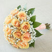 A classic rounded bouquet of apricot-peach roses, arranged with careful symmetry so each blossom nestles into the next, creating an inviting, tactile surface of layered petals. Interspersed among the roses, clusters of white gypsophila breathe lightness into the arrangement, their tiny star-like blooms forming a soft halo that lifts the composition. Broad, glossy green leaves frame and anchor the flowers, their deep colour setting off the warm peach tones and lending an organic, contemporary edge. The stems are bound in a peach ribbon with pearl detailing - a textured, tactile finish that suggests both refined taste and thoughtful care. Bathed in gentle, even daylight, the bouquet's pastel hues and delicate textures read clearly in photographs while remaining true to its natural appearance. The aroma is subtle and inviting: clean green notes of freshly trimmed foliage layered with the sweet, velvety scent of roses and the whisper of baby's breath. This piece is designed with weddings and small celebrations in mind, ideal for a bride who wants a soft, romantic statement or for gifting on an anniversary when you want understated elegance. As someone who arranges flowers for local ceremonies around Exmouth Market and Spa Fields, I picture this bouquet carried through narrow Clerkenwell streets, its compact, cushioned form comfortable in the hand. Clerkenwell Flowers prepares blooms with that neighbourhood attention - careful selection, seasonal freshness, and composition that stands up to the close-up scrutiny of photographs and the emotional weight of the moment.