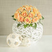 This hand-arranged bouquet captures an elegant simplicity that reads as both contemporary sculpture and heartfelt sentiment across a Clerkenwell mantel or a wedding table on St John Street. A compact dome of peach roses-each bloom unfolding in layered, velvety spirals-creates a warm, honeyed-apricot glow that feels intimate and unforced. Interspersed are clouds of white gypsophila whose tiny, airy blossoms diffuse the light and lend a bridal, ethereal softness, like a delicate veil settling over the roses. The flowers rest within and above a striking, spherical white wicker vessel: an open-weave orb that gives the arrangement an architectural, breathable centre, allowing glimpses of stems and leaves and throwing gentle lattice shadows on surrounding surfaces. Greenery is used sparingly but with purpose, the cool, matte leaves providing contrast and framing the rose heads so they read as a single, harmonious dome. Light plays a quiet role here-soft daylight from a Clerkenwell window warms the peach tones and edges the petals with a faint golden rim, while the overall scent is a tender, floral whisper-rose powder softened by fresh green notes. As a florist working around Leather Lane and the Barbican, I arrange these pieces to feel right in both modern flats and historic brick-lined venues; they suit intimate weddings, milestone dinners, or a calm, considered gift. Clerkenwell Flowers prepares this style with attention to balance and proportion and can craft similar pieces for local delivery within the area.